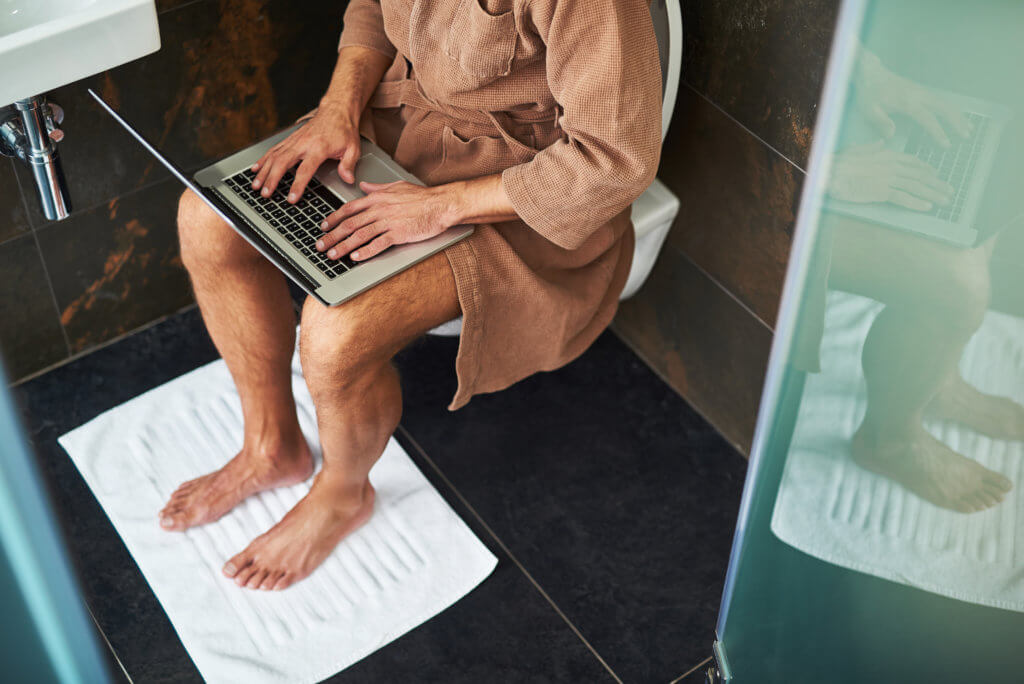 Man in bathrobe using laptop while sitting on toilet at home