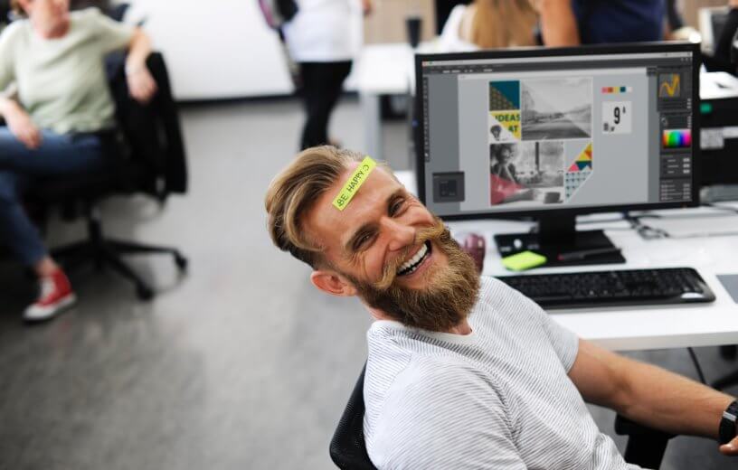 Office employee with "Be Happy" sticker on his head while sitting at desk