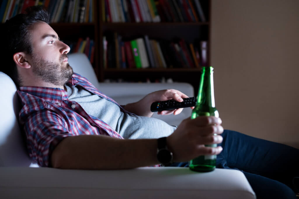 Bored man alone watching TV, drinking beer on couch