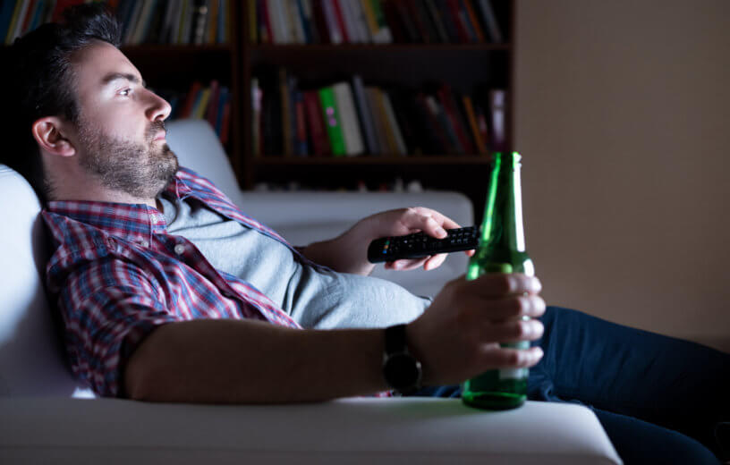 Bored man alone watching TV, drinking beer on couch