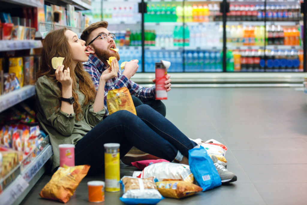 Couple sitting on supermarket floor eating chips, snacks