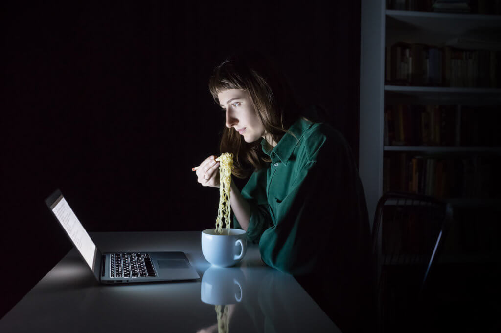 Woman eating noodles as late night dinner in front of computer in dark room