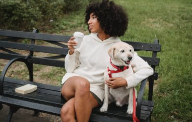Woman sitting outside on park bench with dog while drinking coffee