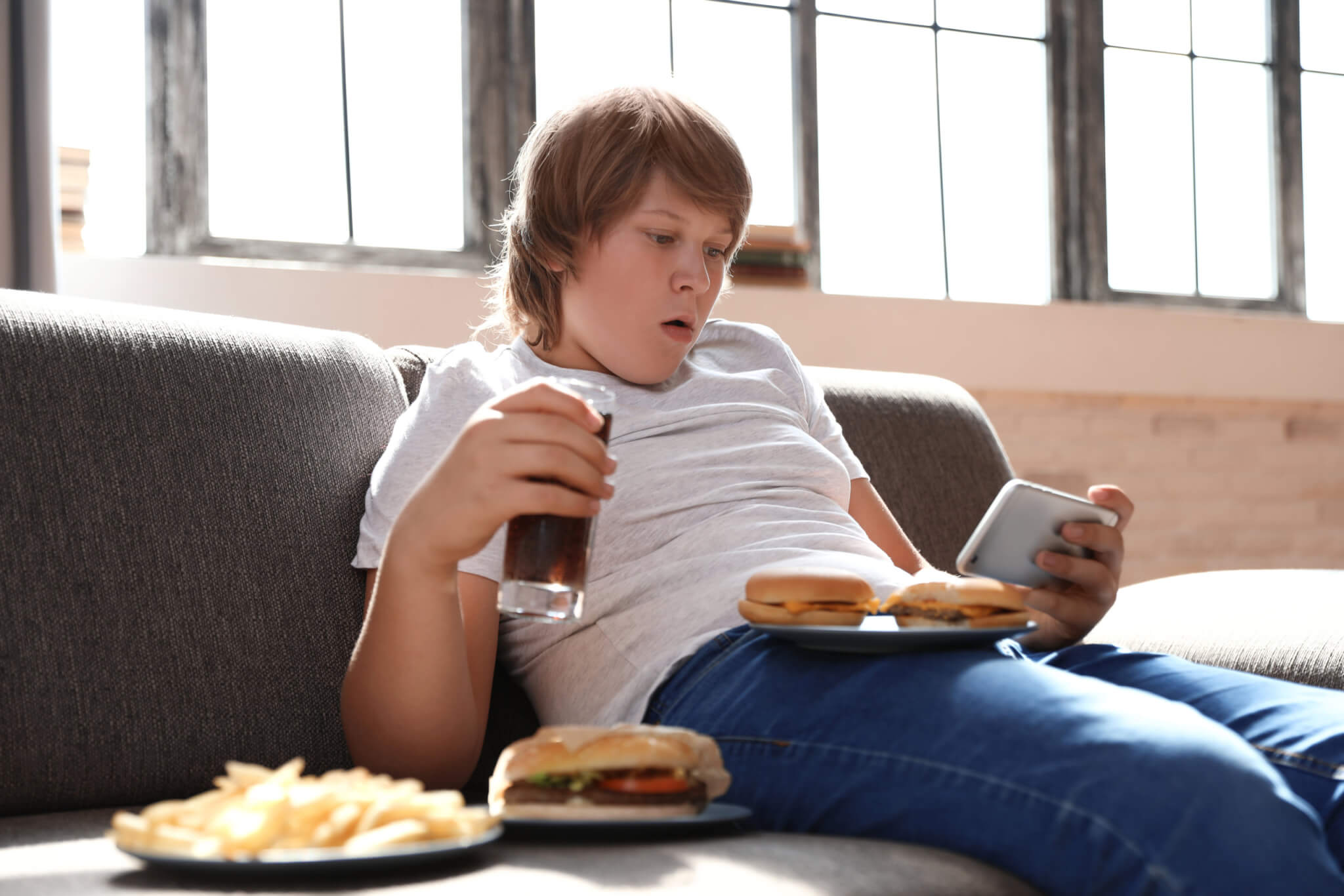 Teen boy eating junk food, drinking soda while looking at smartphone