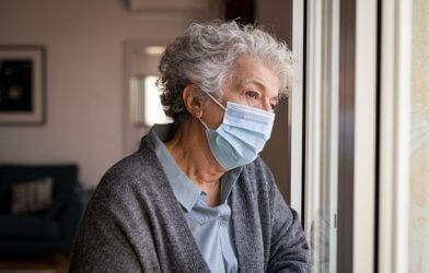 Older woman wearing face mask looking out window, sad, alone during COVID pandemic