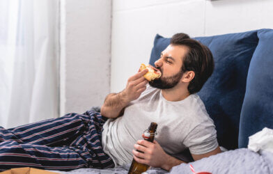 Man eating pizza and drinking beer alone in bed