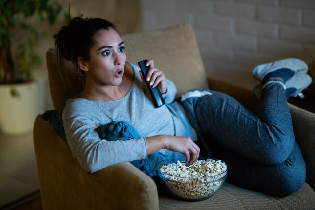 Young shocked woman watching movie on TV and eating popcorn at night.