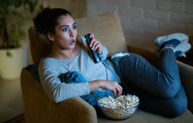 Young shocked woman watching movie on TV and eating popcorn at night.