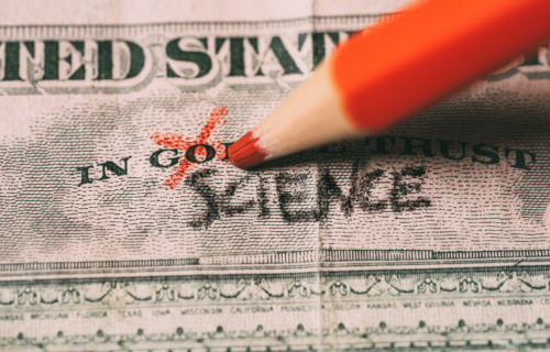 Hand crossing out the word "God" for science on on a US dollar bill