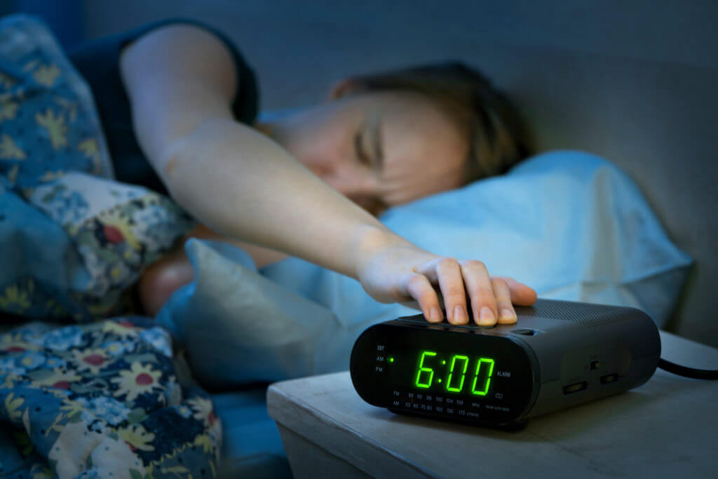 Woman hitting snooze button on alarm clock after waking up so she can sleep longer