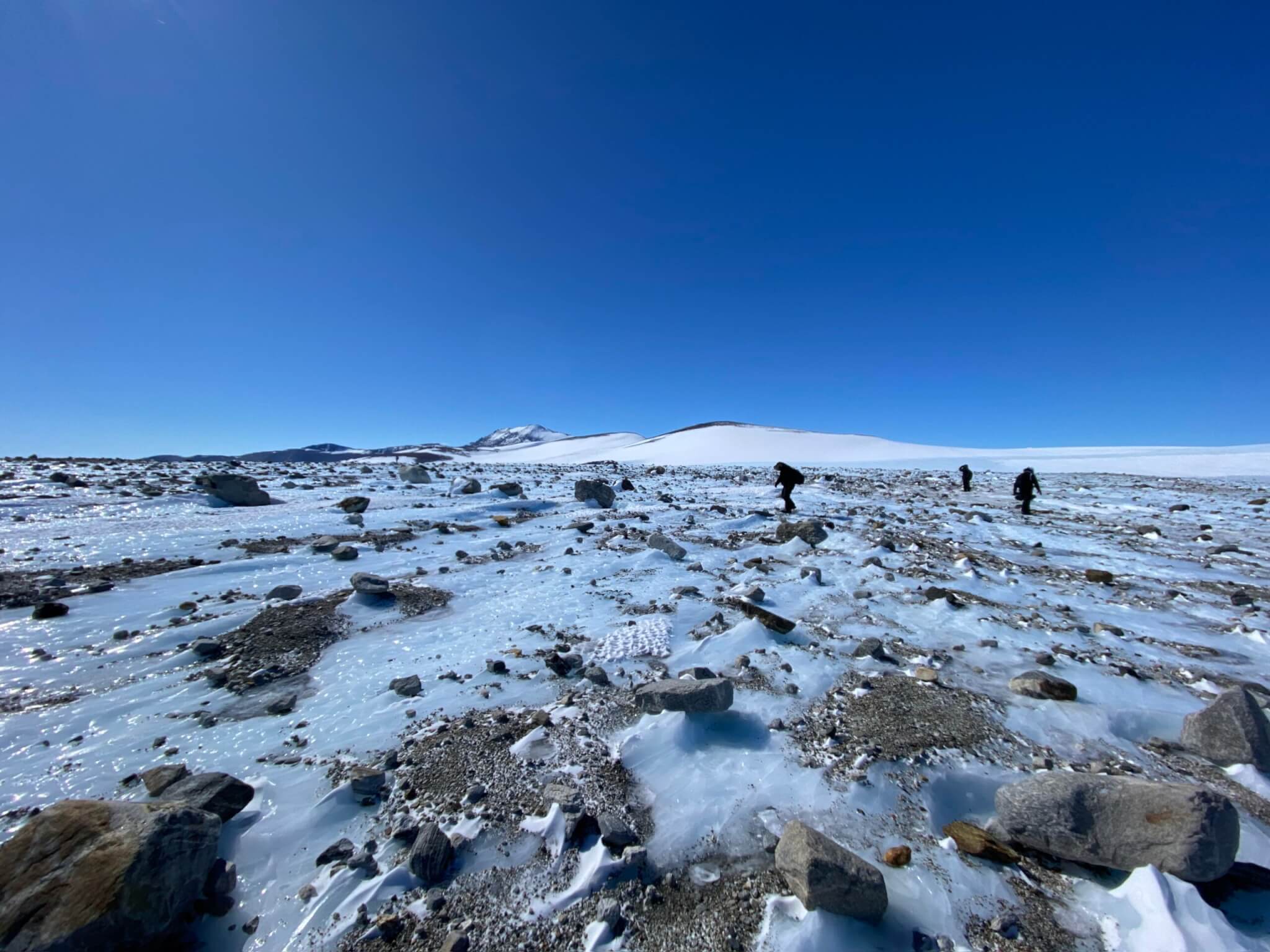 Rocks strewn across an ice field, with the scientists searching for meteorites in the background.