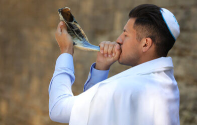 Jewish man in kippah and tallit blowing shofar outdoors to celebrate Rosh Hashanah