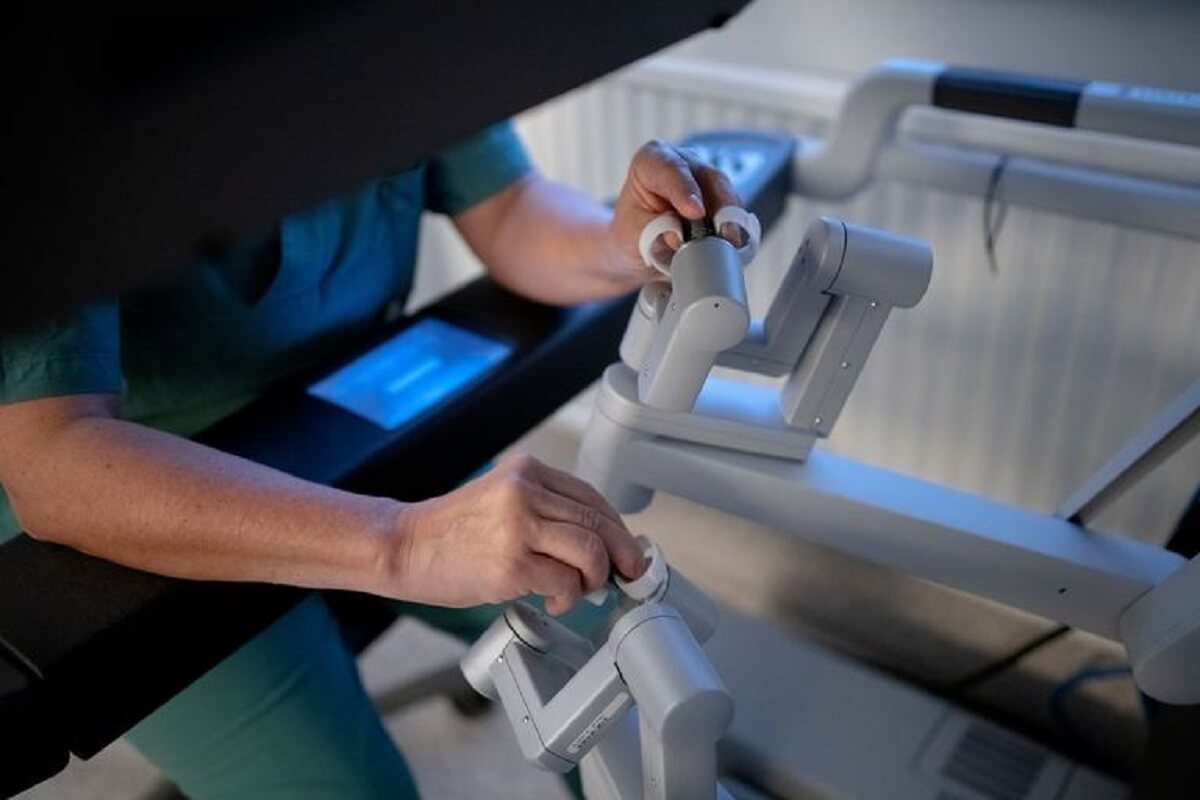 close of a surgeon's arms and hands, doctor is wearing green scrubs. He is touching knobs on two gray robotic arms 