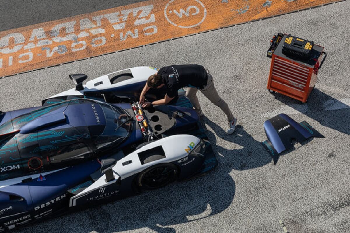 close aerial view of dark blue racecar with white stripes along the front of the body, parked on a race track , a man is behind over the car checking the engine. behind him is an orange toolbox