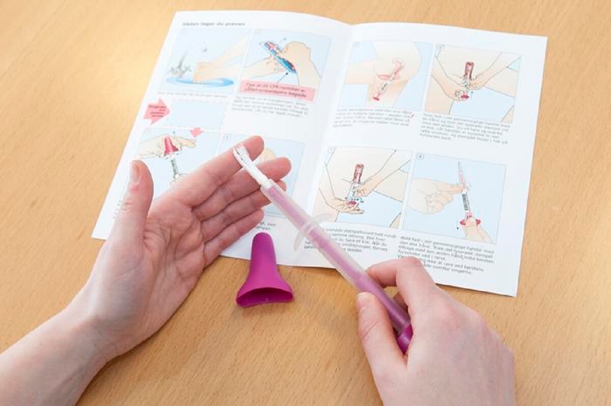 person holding a white plastic stick with hands.hands are hovering over a pamhlet show different steps of taking an hpv test