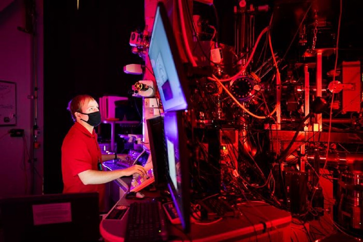 researcher wearing black mask sits in front several monitors in a room with red lighting, uses microscope to study fatigue cracks in materials