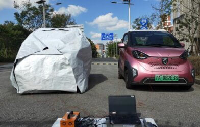 two cars in a parking lot, once covered by a thermal cloak, the other is a red SUV uncovered. In front the cars are devices that measure temperatures