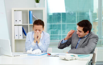Man covers his face while his sick coworker blows her nose at the office.