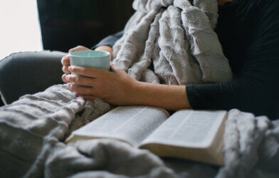 Woman drinking coffee and reading a book under a blanket