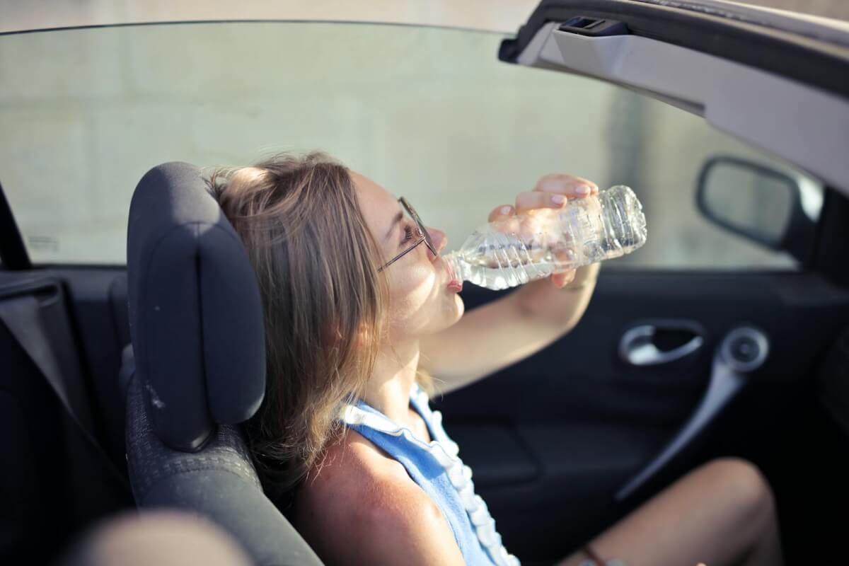 High angle side view of young woman in glasses and casual clothes drinking water from plastic transparent bottle while sitting in cabriolet with open roof in traffic jam in hot sunny day