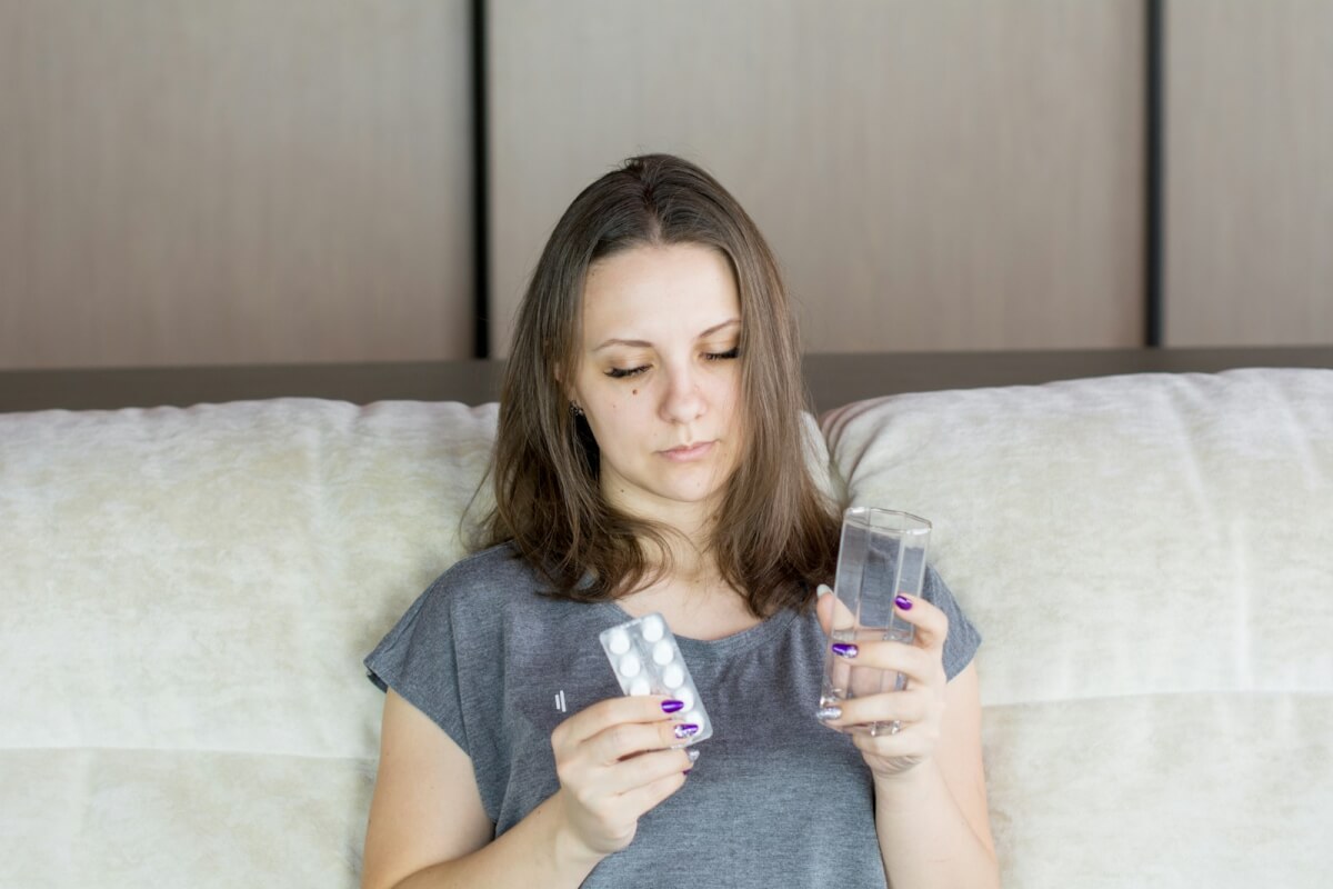 a woman sitting on a couch looking at a pack of pills and holding a glass of water